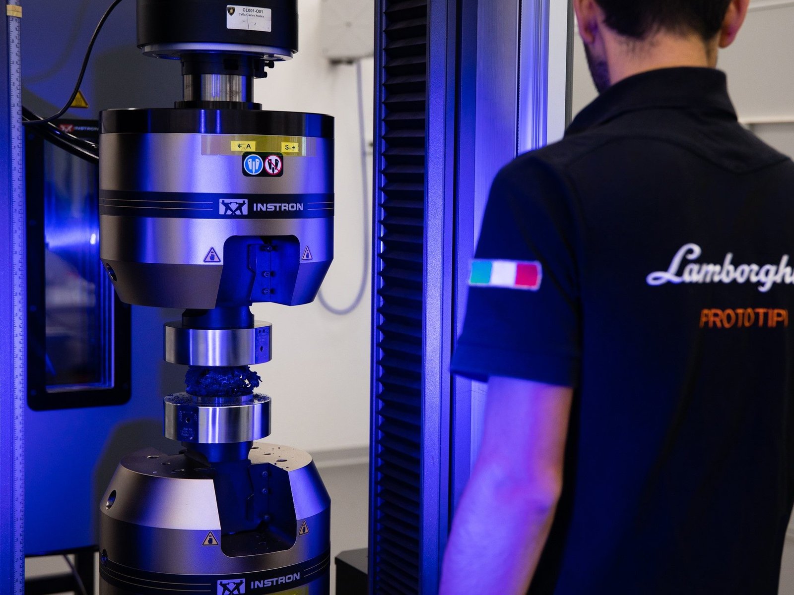 Lamborghini engineer monitoring a carbon fiber compression test on an Instron machine under blue UV lighting at the ACLSD laboratory