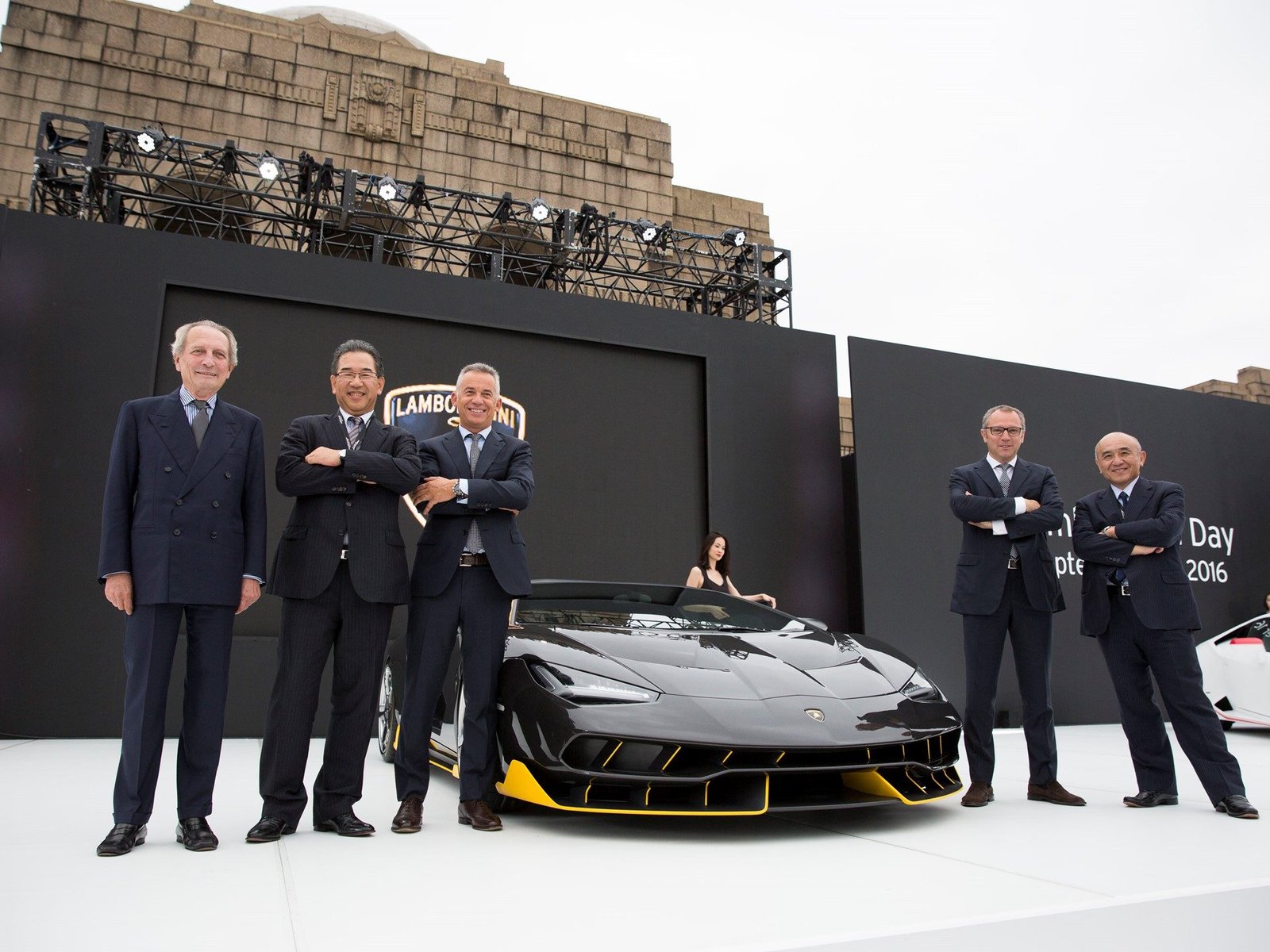 Black Lamborghini Centenario Roadster with yellow accents displayed at the Excellence in Carbon Fiber event in Tokyo, with executives surrounding the car