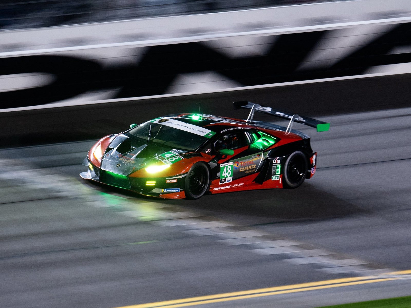 The #48 Paul Miller Racing Lamborghini Huracán GT3 Evo carving through the Daytona banking at night with green and yellow lighting visible