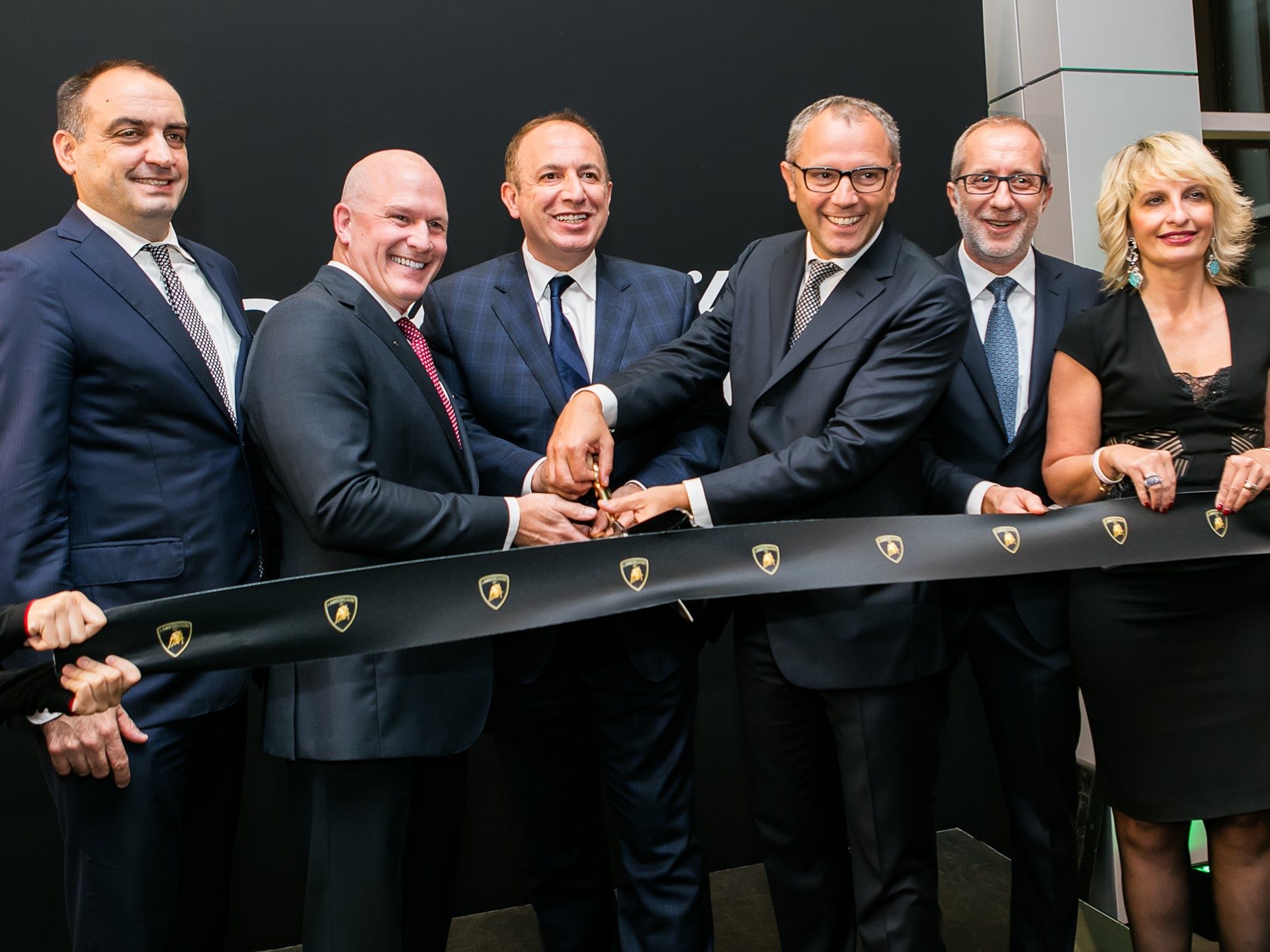 Executives and VIPs cutting a black ribbon with gold Lamborghini logos at a dealership grand opening ceremony