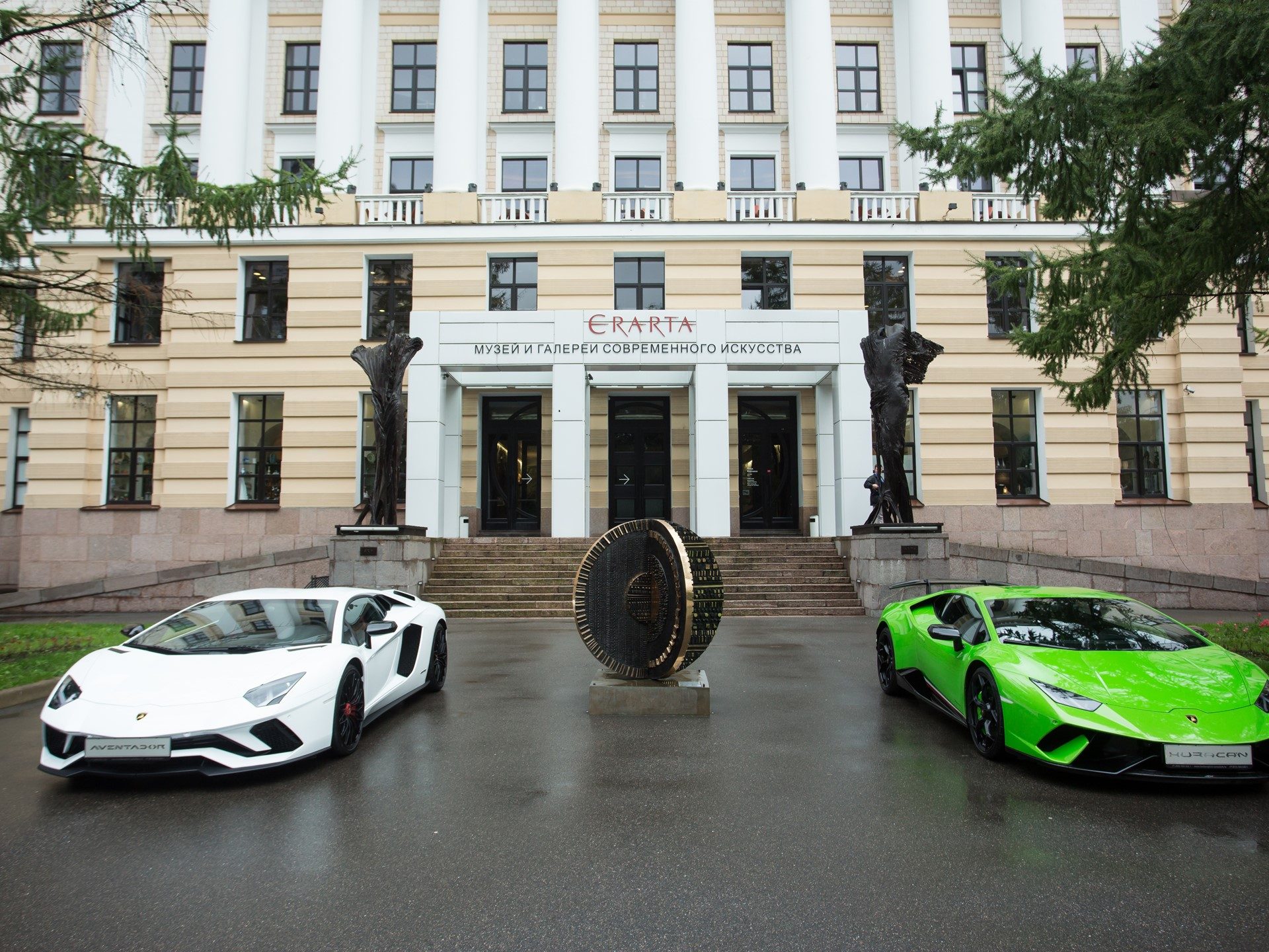 A white Lamborghini Aventador and green Huracan parked before a grand columned building in St. Petersburg