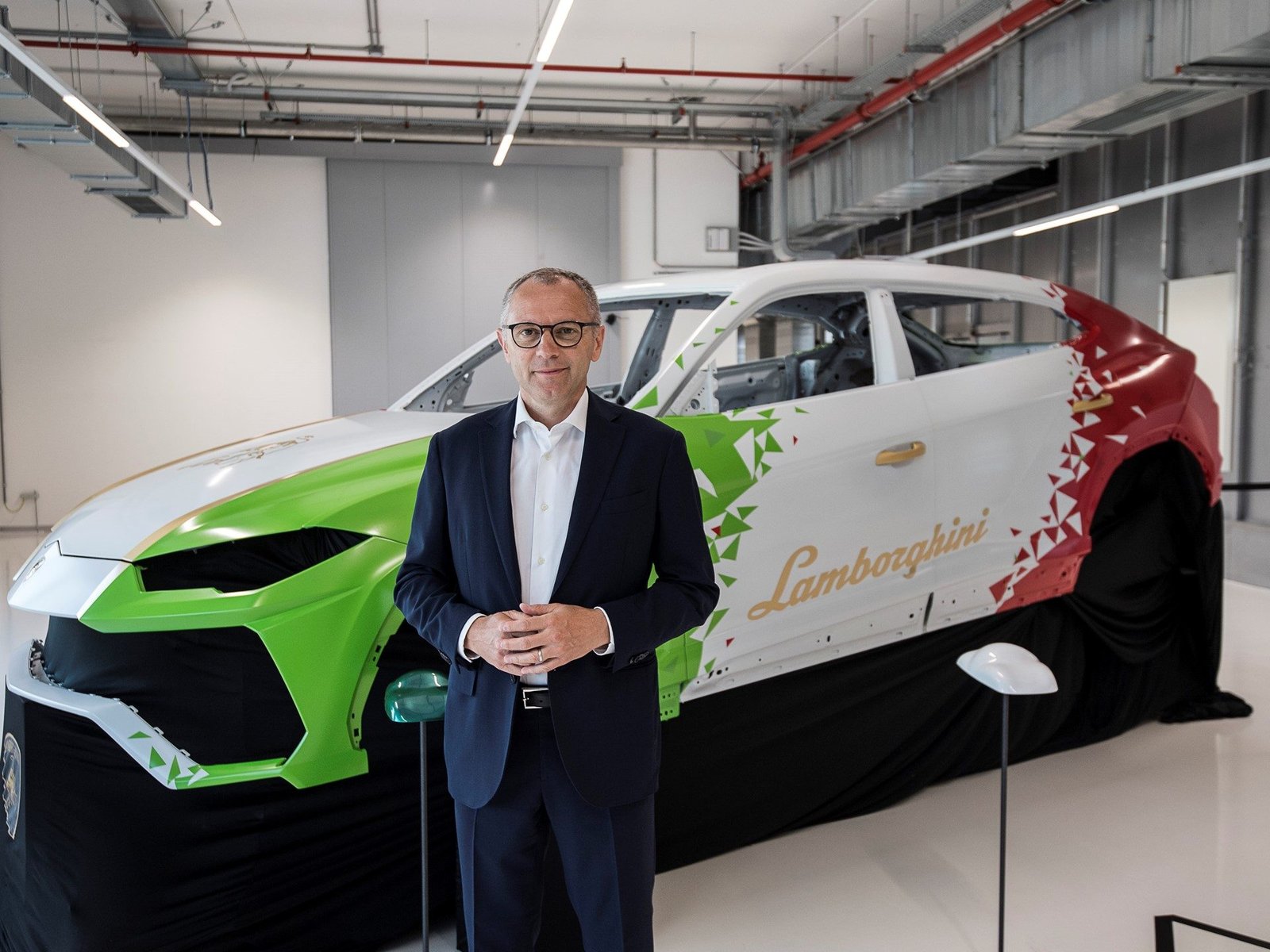 A Lamborghini executive stands beside a partially assembled Urus body shell painted in Italian tricolor livery inside the Sant'Agata Bolognese factory