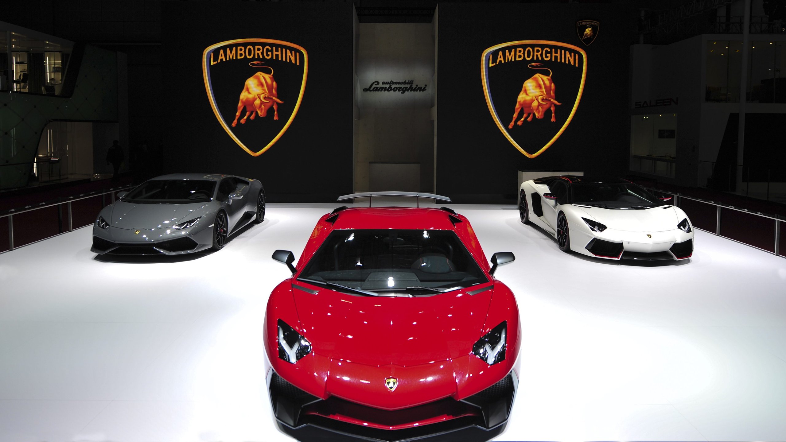 Three Lamborghini supercars displayed on a white platform at Auto Shanghai 2015, including a red Aventador, grey Huracán, and white Aventador under large Lamborghini branding