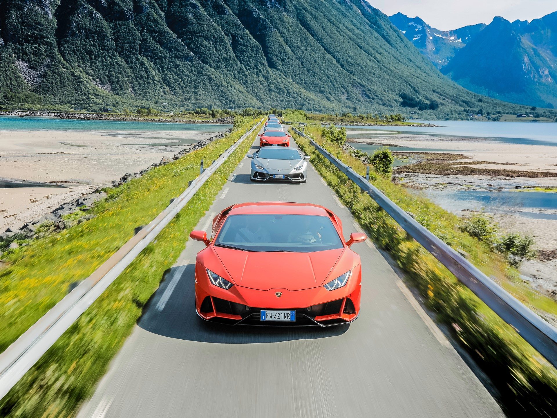Line of Lamborghini Huracán EVO supercars driving through the Lofoten Islands with Y-shaped LED running lights visible on the lead orange car