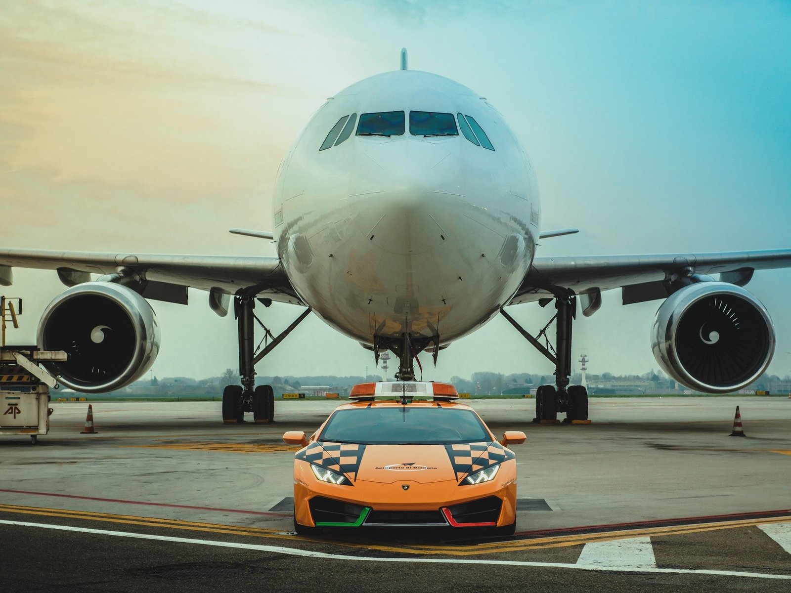 Orange Lamborghini Huracán RWD Follow-Me car with checkered hood and roof-mounted light bar parked in front of a commercial airliner at Bologna Airport