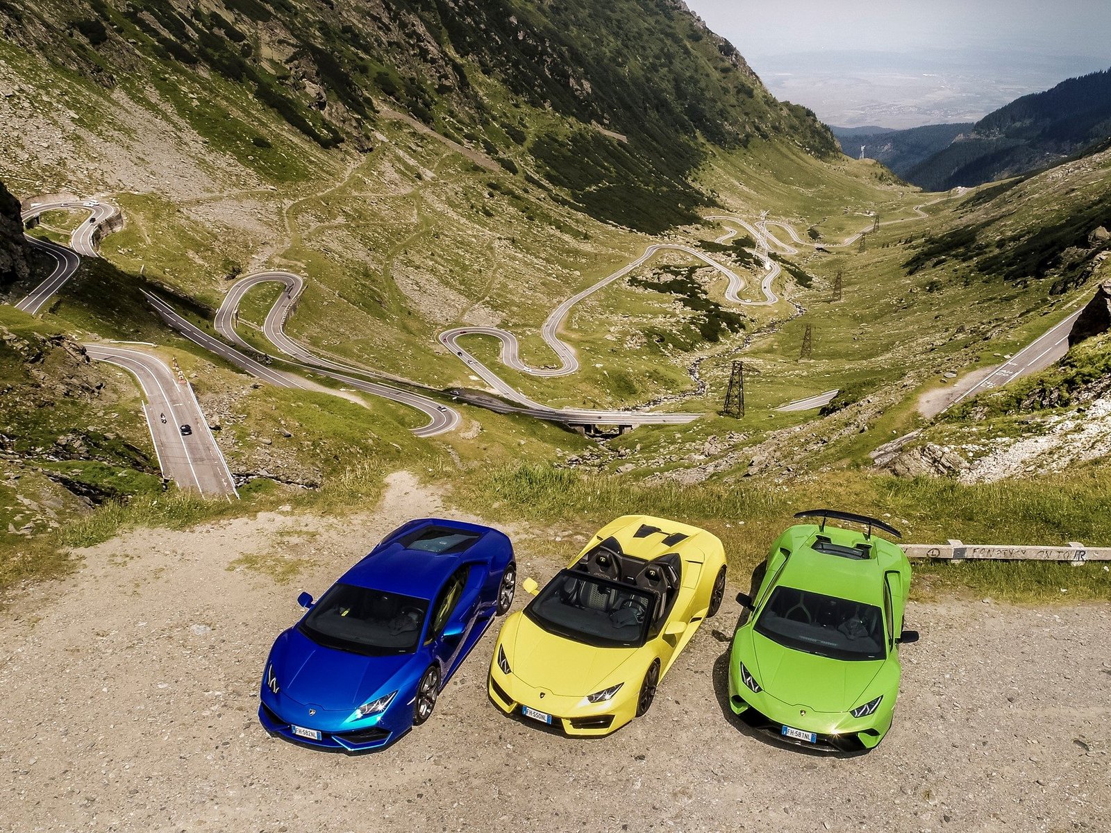 Aerial view of three Lamborghini Huracáns in blue, yellow, and green parked on a dirt pull-off overlooking the winding Transfăgărășan mountain road in Romania
