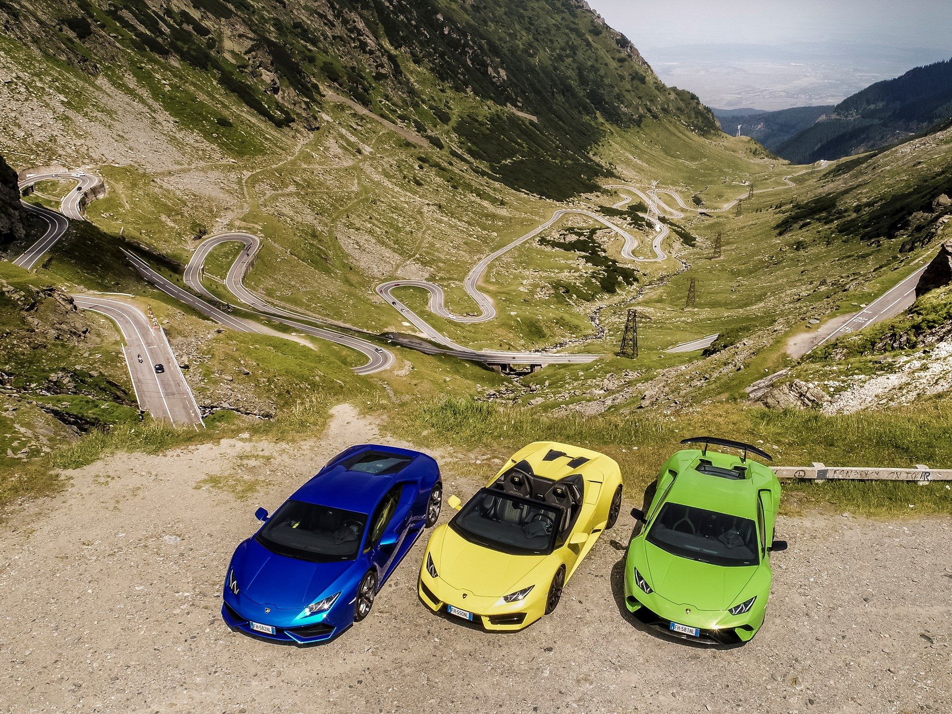Aerial view of three Lamborghini Huracáns in blue, yellow, and green parked on a dirt pull-off overlooking the winding Transfăgărășan mountain road in Romania