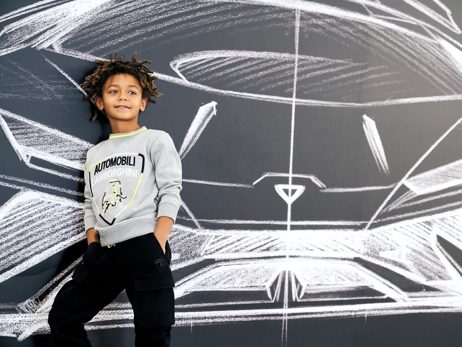 Child model in grey Lamborghini sweatshirt standing before chalk car drawings on a dark wall