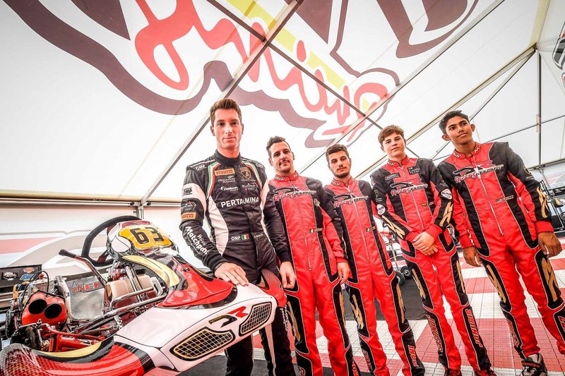 Kart Drivers Program participants in red and black racing suits standing with a DR Racing Kart under a branded tent at Adria