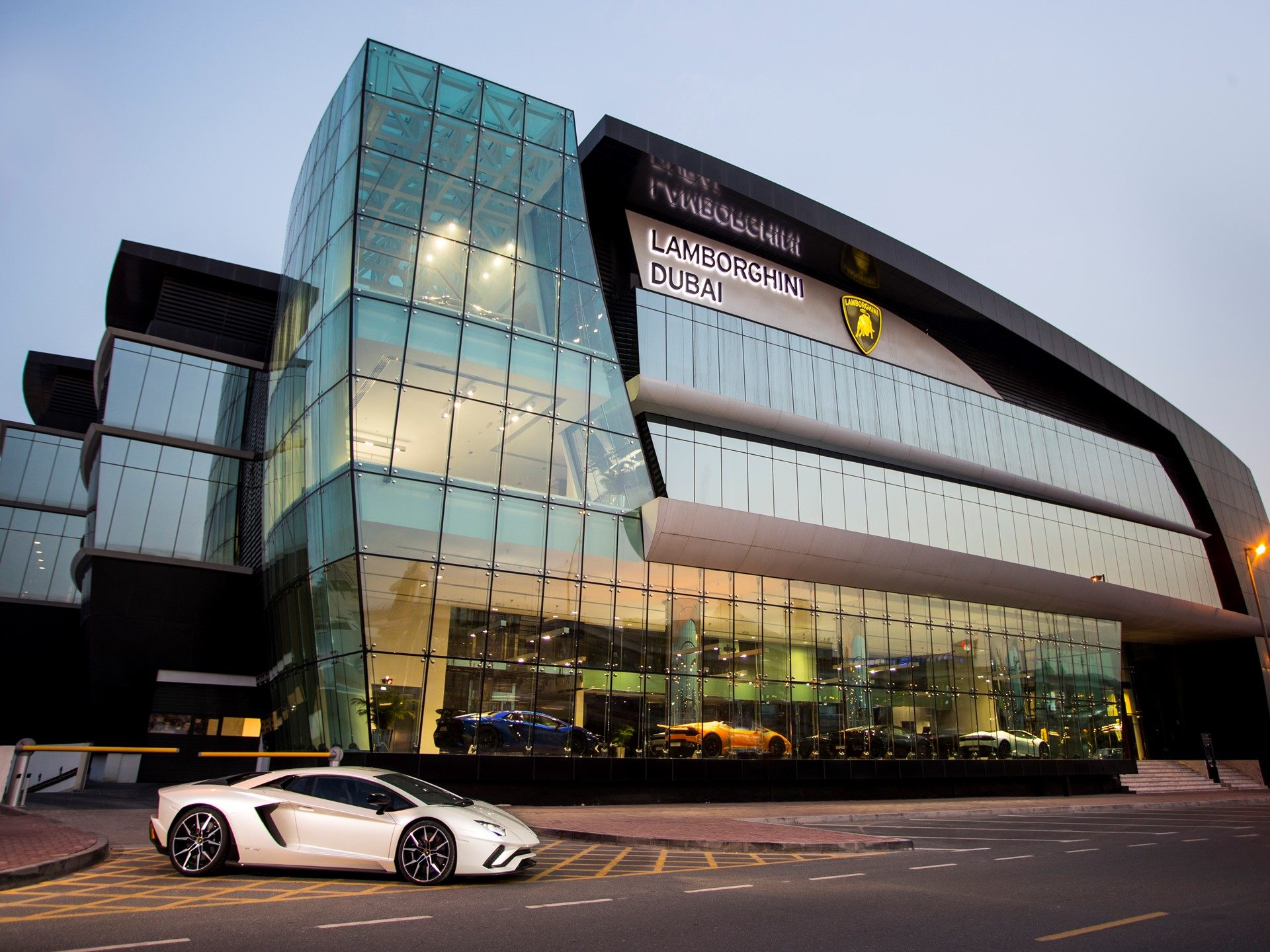White Lamborghini Aventador parked outside the glass-fronted Lamborghini Dubai showroom on Sheikh Zayed Road, with colorful models visible inside