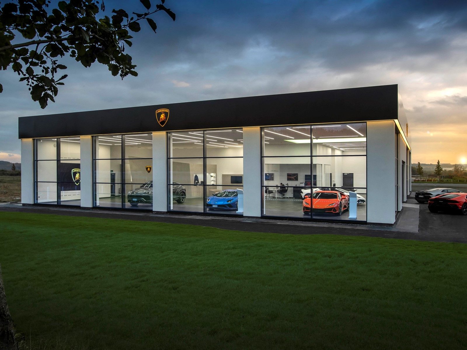 Lamborghini Leeds showroom exterior at dusk with a green Urus, blue Huracán, and orange Huracán visible through the glass facade