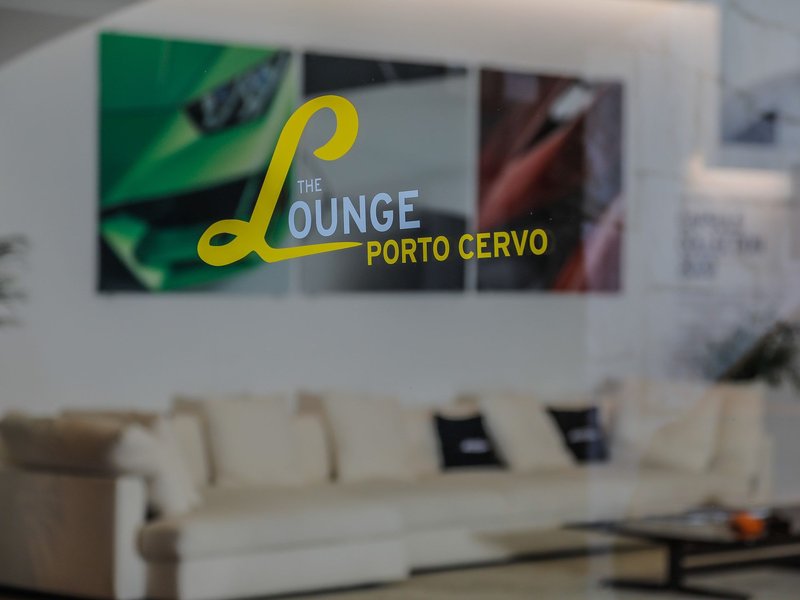 The Lounge Porto Cervo logo etched on a glass partition inside the Lamborghini Lounge, with a white sofa and abstract car imagery visible in the background