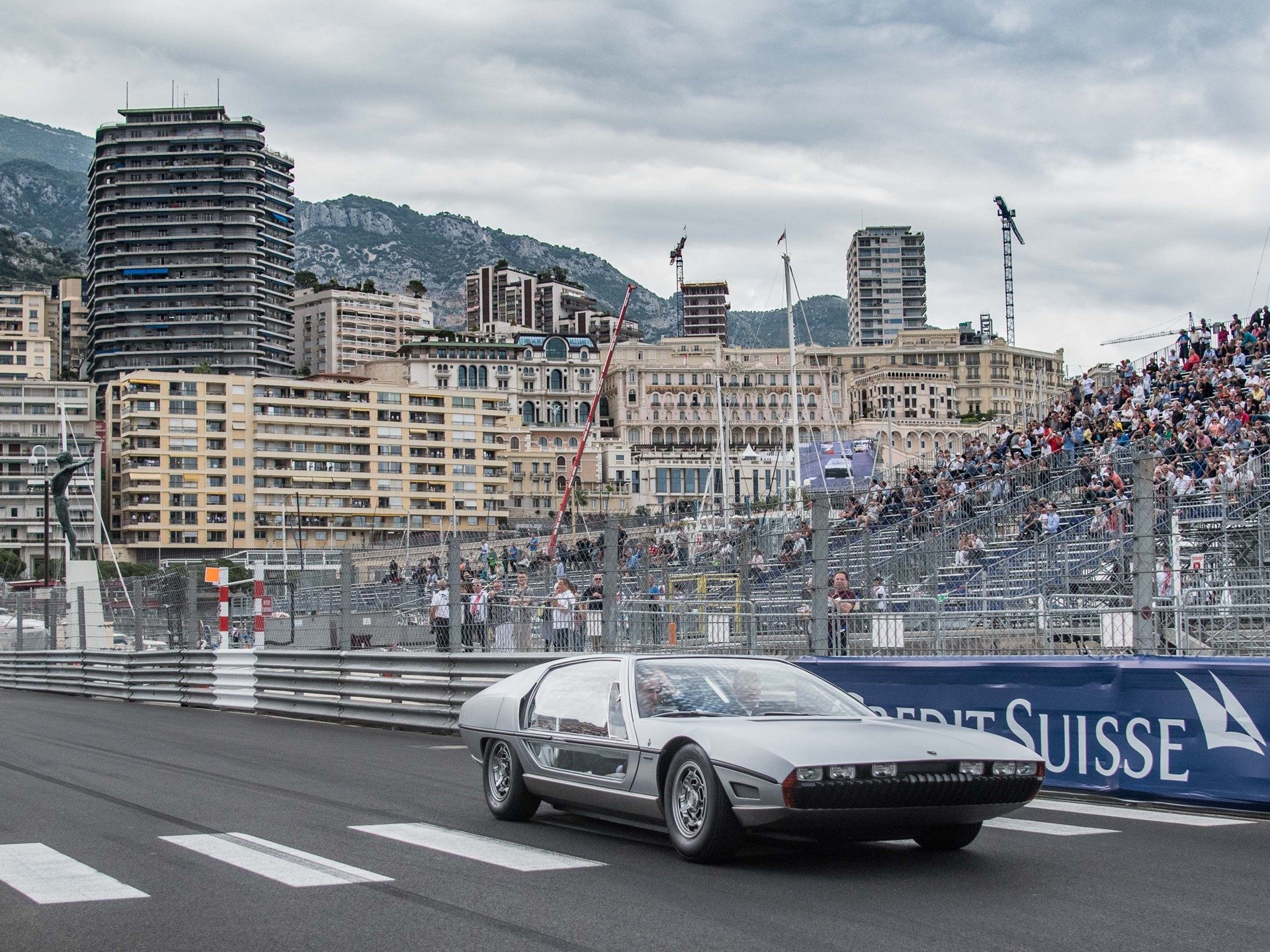 Lamborghini marzal concept car in silver driving on the monaco circuit under cloudy skies with spectators visible in grandstands