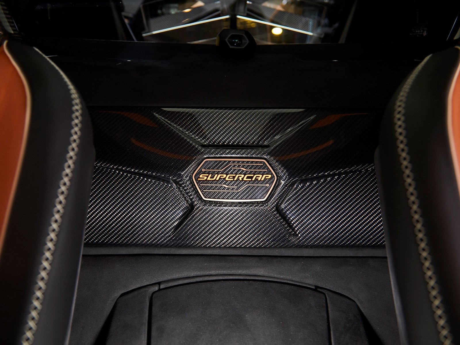 Close-up of a carbon fiber panel with bronze SUPERCAP emblem flanked by brown leather seats with white stitching