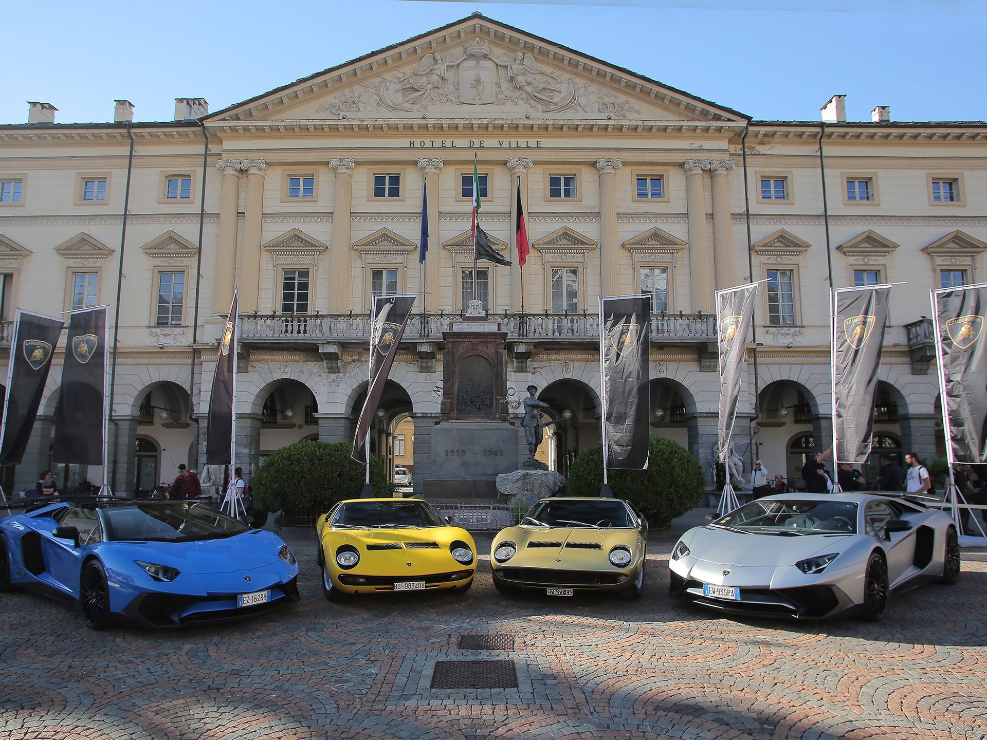 Two Lamborghini Miuras and two Aventadors parked in a cobblestone European square with Lamborghini flags and a grand Hotel de Ville building in the background