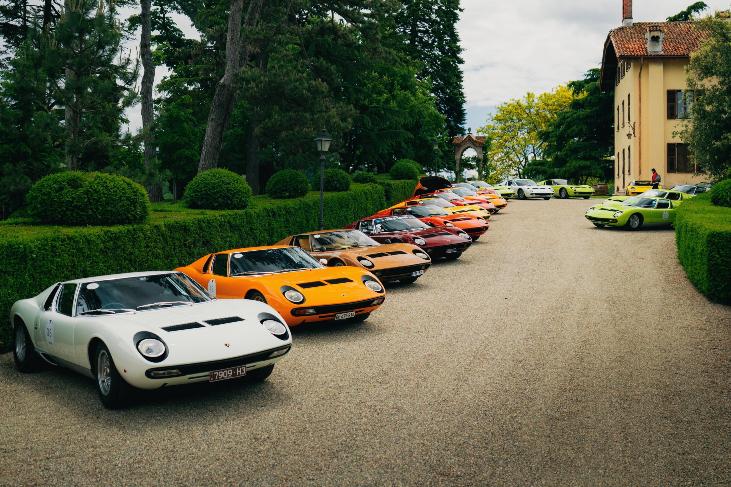 A long line of lamborghini miuras in white, orange, red, and green parked on a gravel driveway leading to a historic building