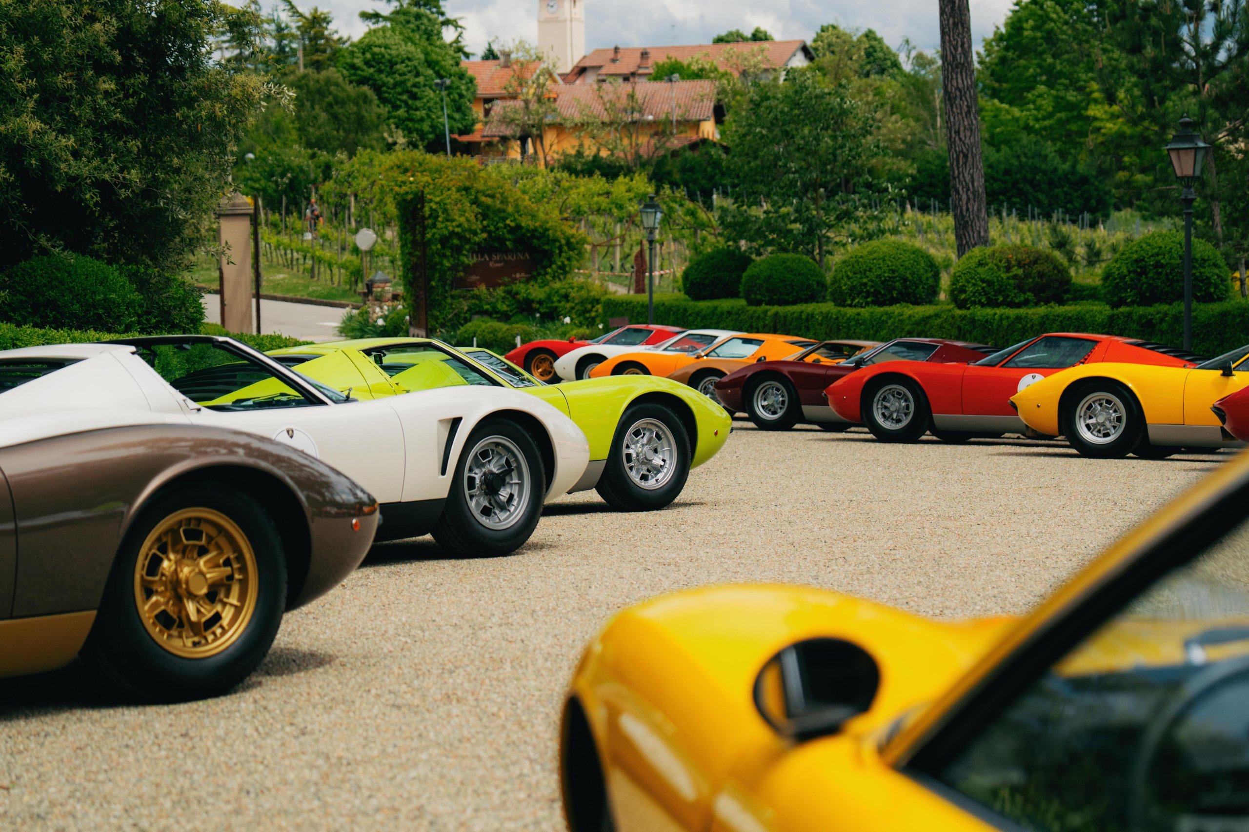 A yellow lamborghini miura in the foreground with a line of colorful miuras in brown, white, green, red, and orange behind it on a gravel driveway