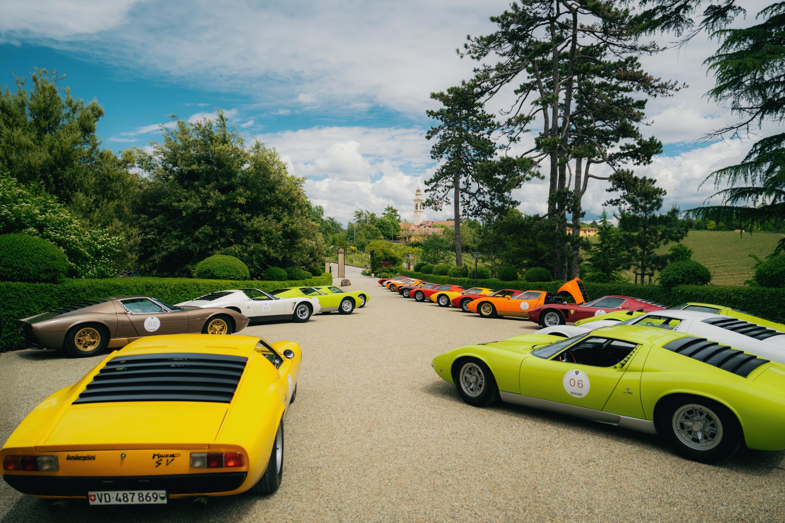 A large collection of lamborghini miuras in yellow, green, white, brown, orange, and red parked on a gravel driveway in a scenic outdoor setting