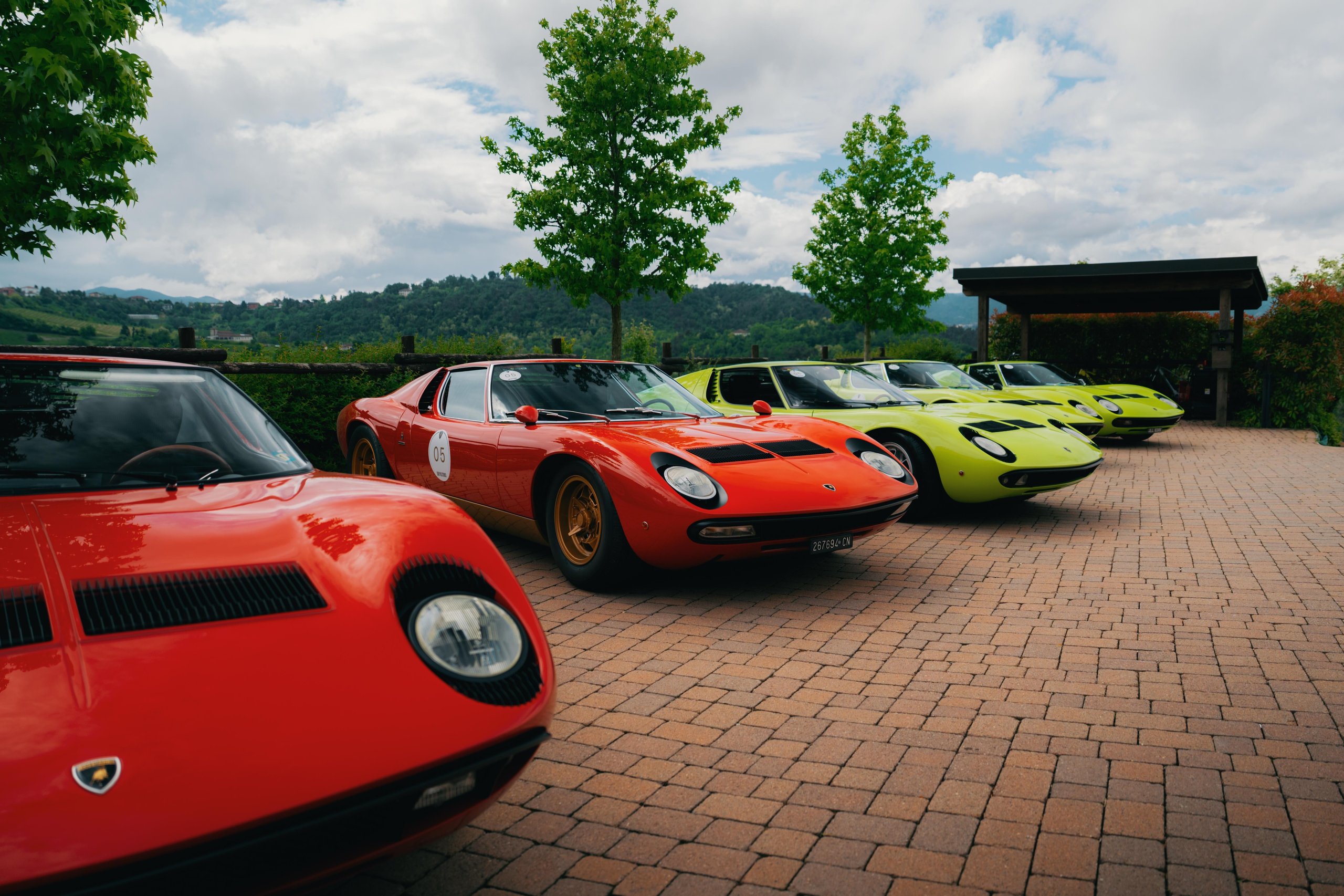 A line of colorful lamborghini miuras including red, orange, and lime green parked on a brick driveway with trees and hills behind