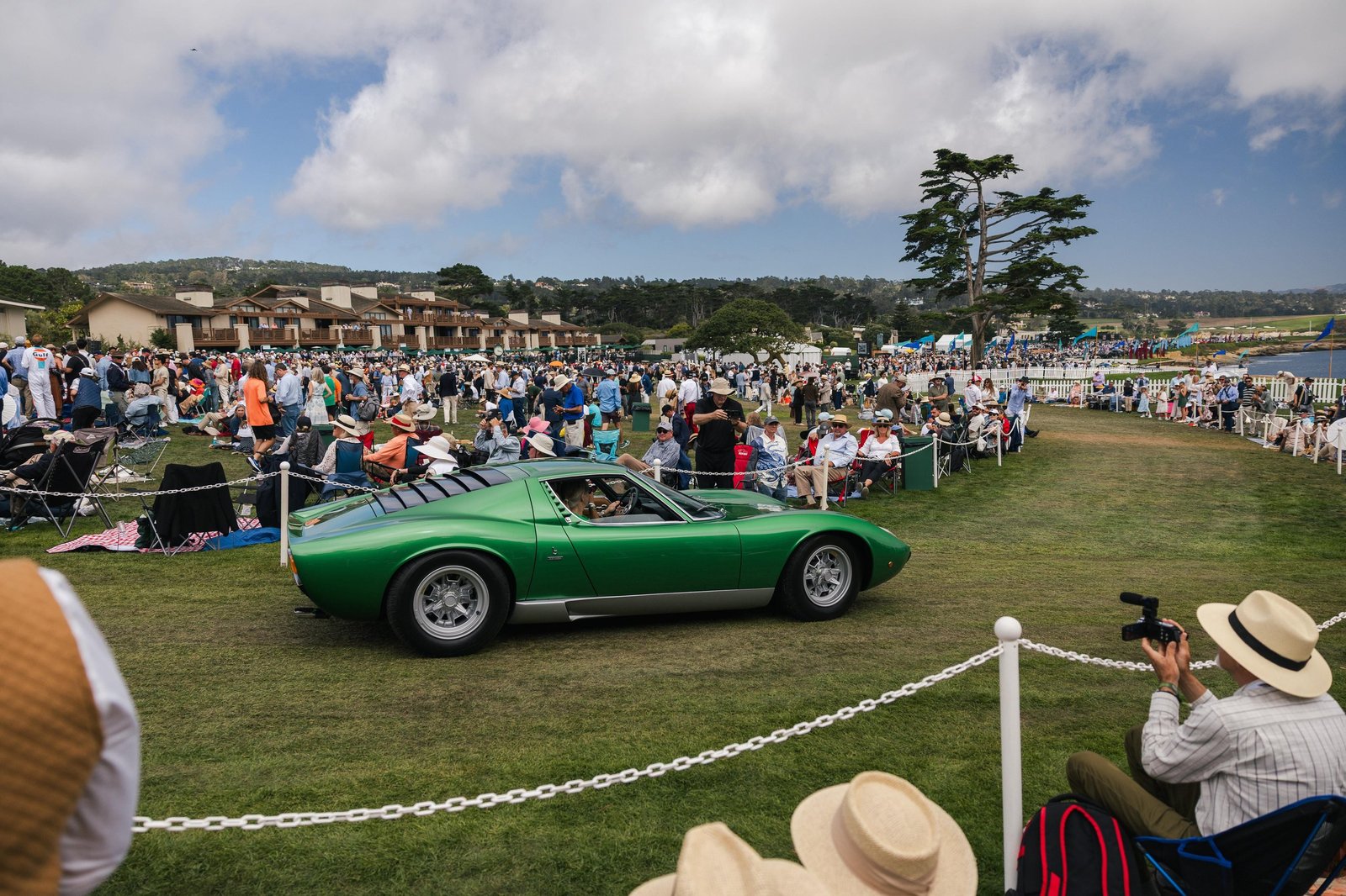 A green Lamborghini Miura driving on a grassy field at an outdoor concours event with spectators in the background