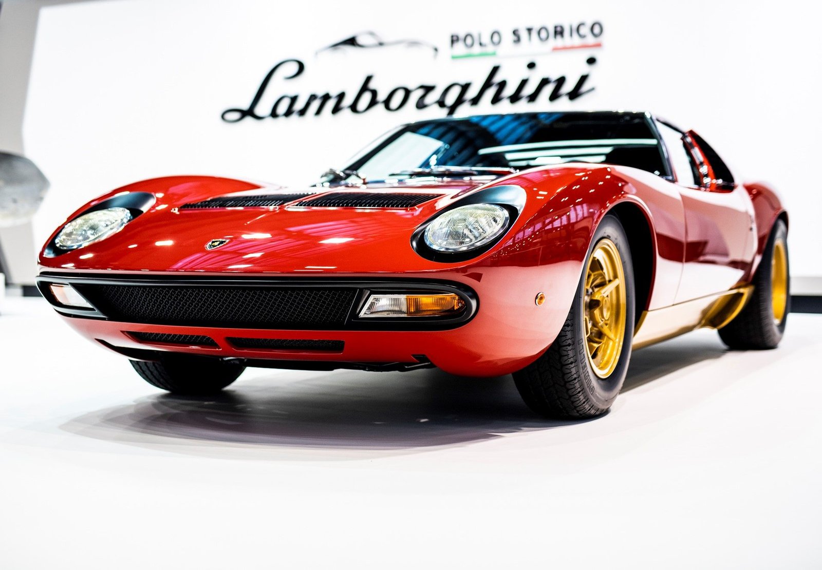 Classic red Lamborghini Miura P400 photographed from a low front angle showing pop-up headlights, gold Campagnolo wheels, and Bertone styling, with Lamborghini Polo Storico logo in background