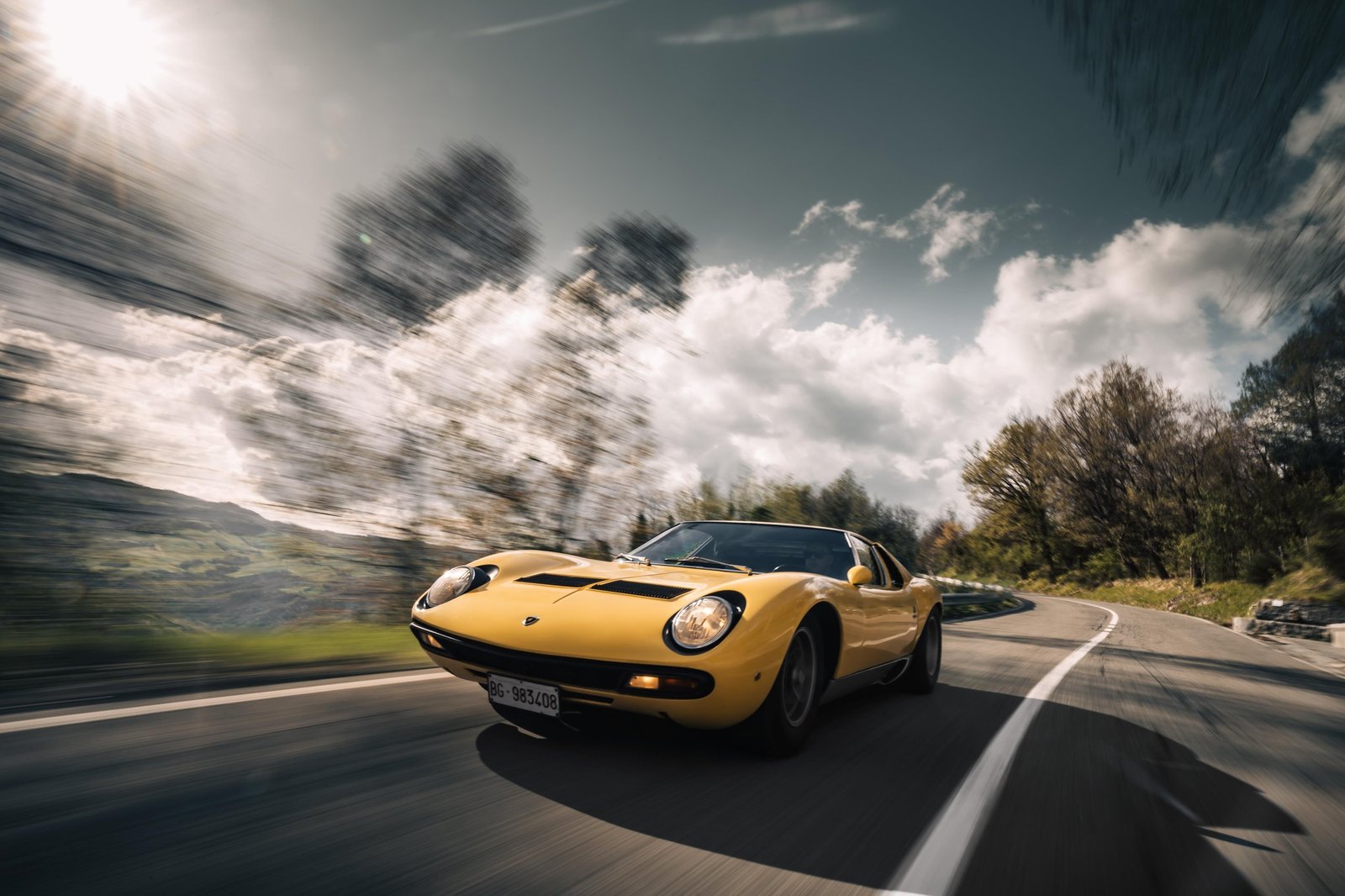 Yellow Lamborghini Miura SV speeding along a winding mountain road with motion blur emphasizing velocity