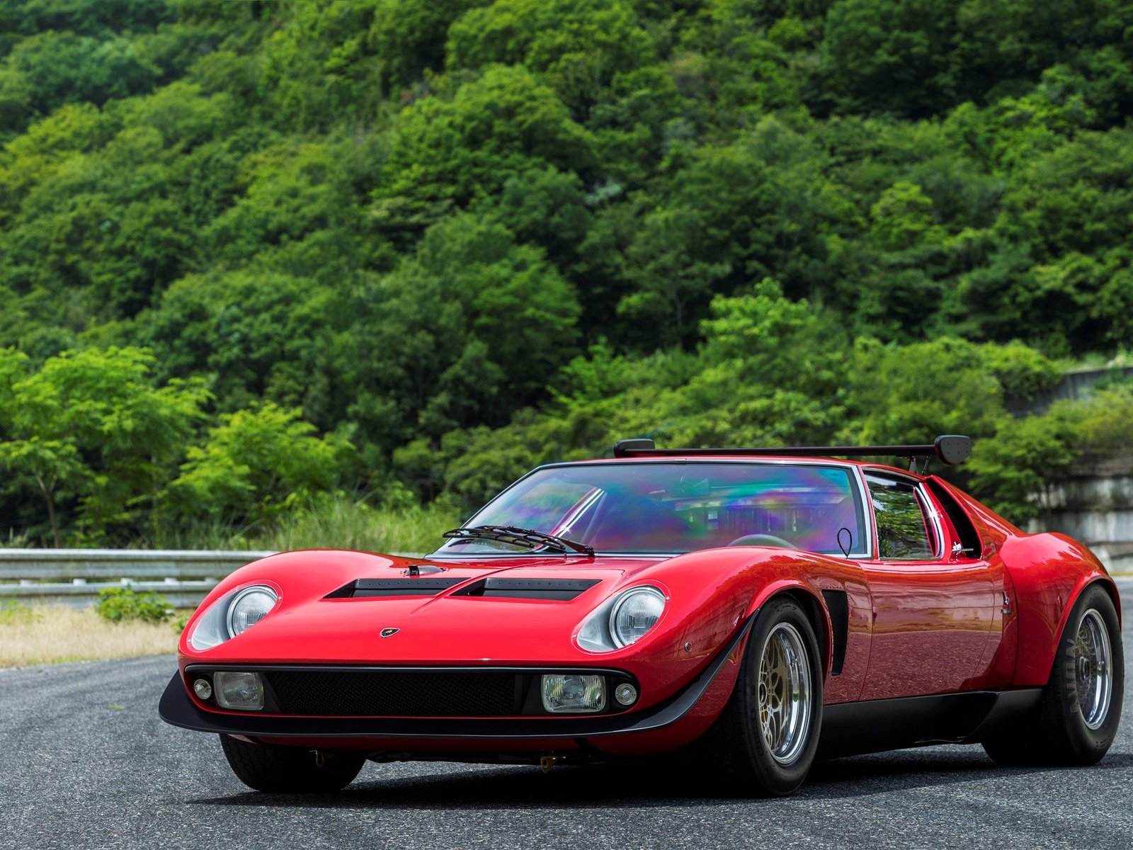 Red Lamborghini Miura SVR showing its wide stance, rear spoiler, and pop-up headlights against a green hillside