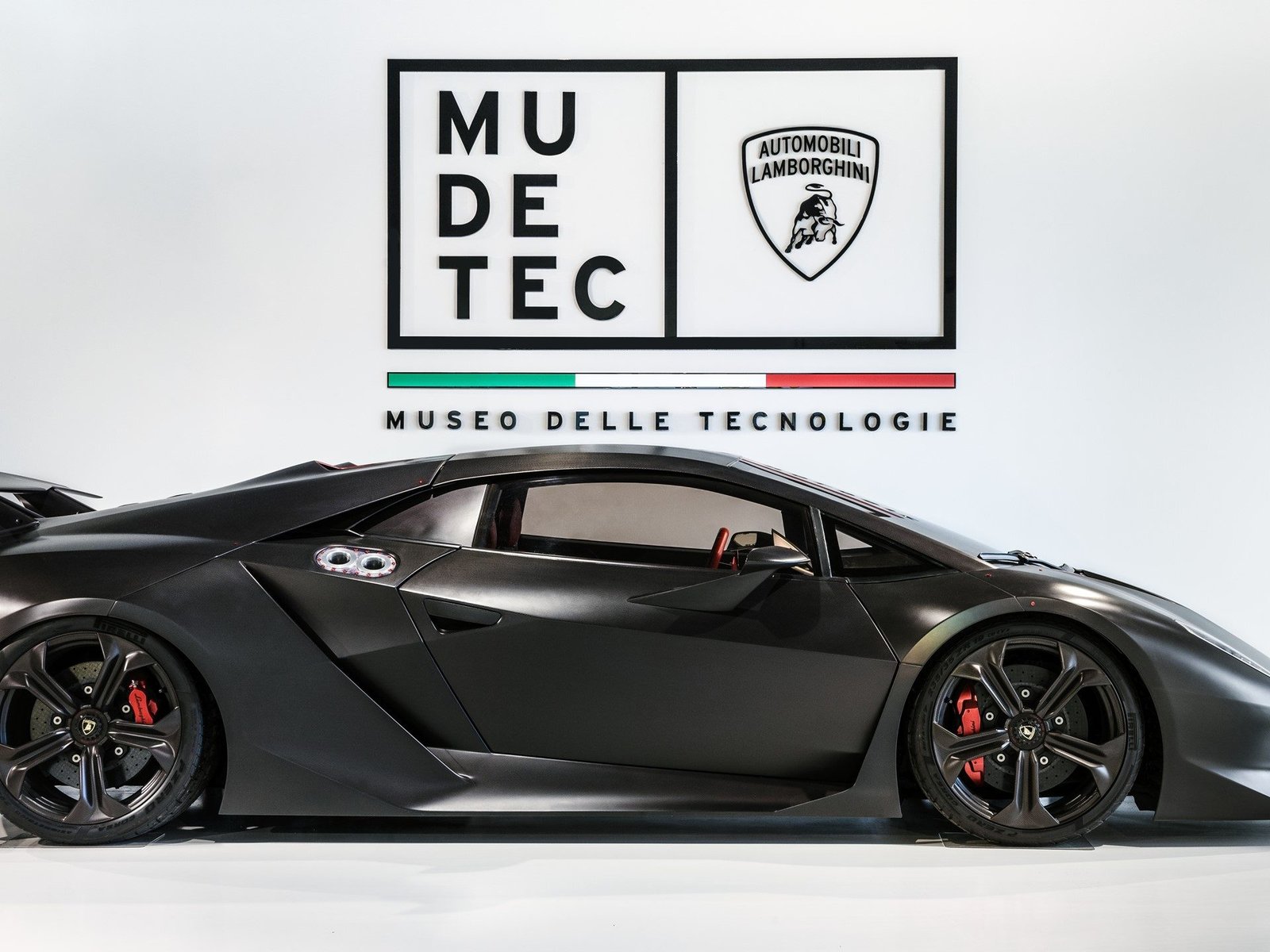 Lamborghini Sesto Elemento in matte black carbon fiber displayed at MUDETEC with the museum's branding visible on the wall behind