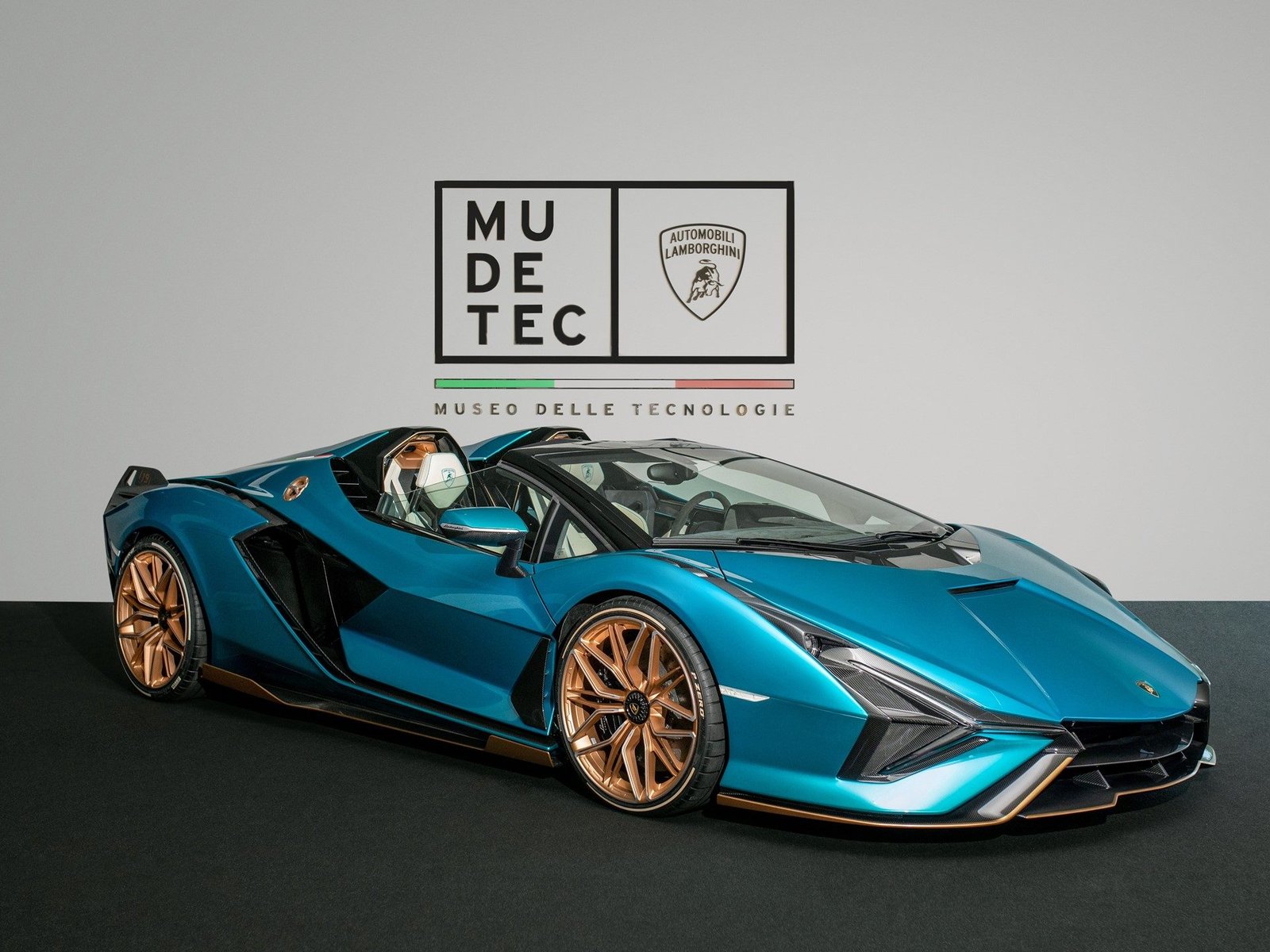 Teal Lamborghini Sián Roadster on display inside the MUDETEC museum in Sant'Agata Bolognese, with MUDETEC and Automobili Lamborghini logos visible on the wall behind it