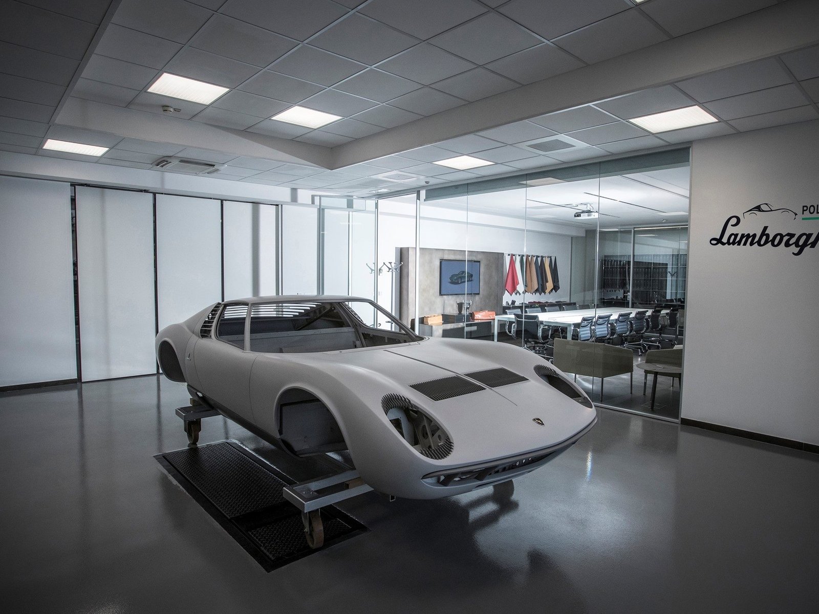 Stripped Lamborghini Miura body shell on a dolly inside the Polo Storico workshop, with a conference room visible through glass walls