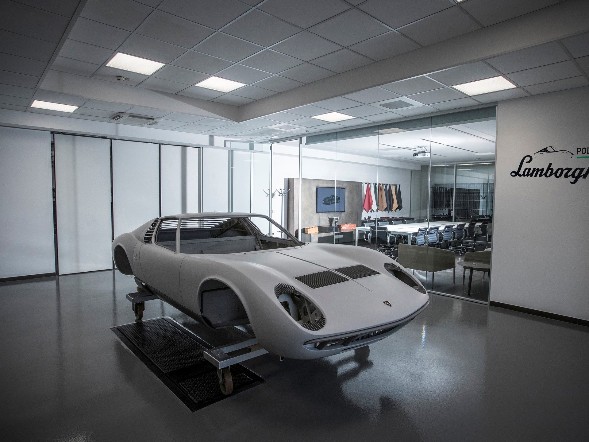 Stripped Lamborghini Miura body shell on a dolly inside the Polo Storico workshop, with a conference room visible through glass walls