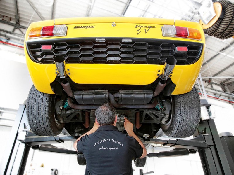 A yellow Lamborghini Miura SV on a lift at the Polo Storico facility, with a technician in an Assistenza Tecnica Lamborghini shirt working underneath