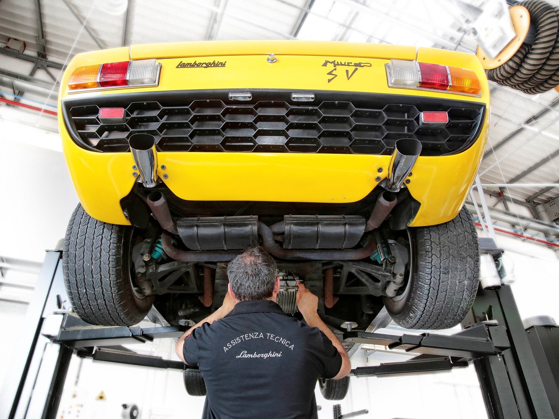 A yellow Lamborghini Miura SV on a lift at the Polo Storico facility, with a technician in an Assistenza Tecnica Lamborghini shirt working underneath