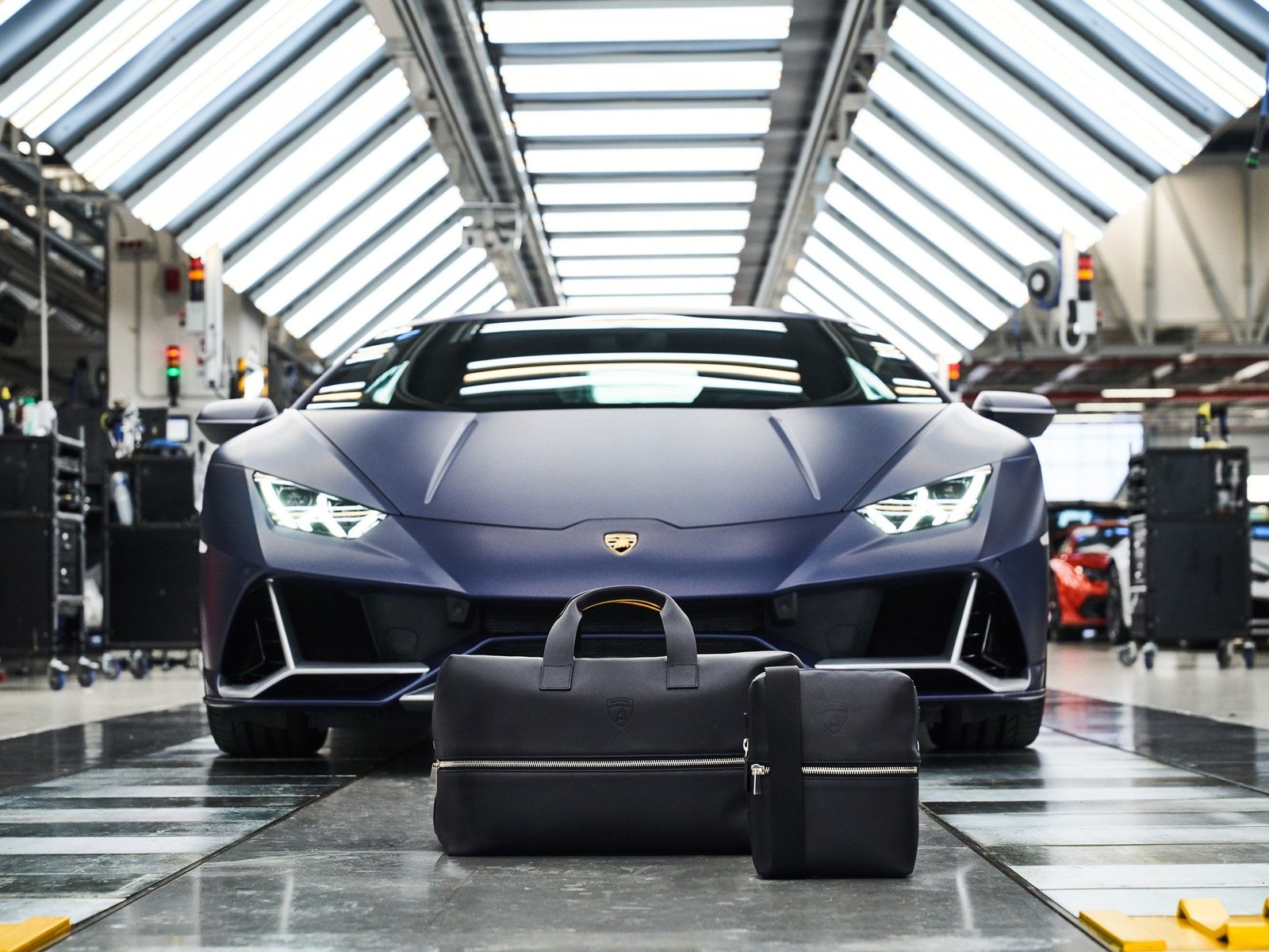 Two black leather bags from the Automobili Lamborghini collection by Principe displayed in front of a matte blue Huracán Tecnica in a factory setting