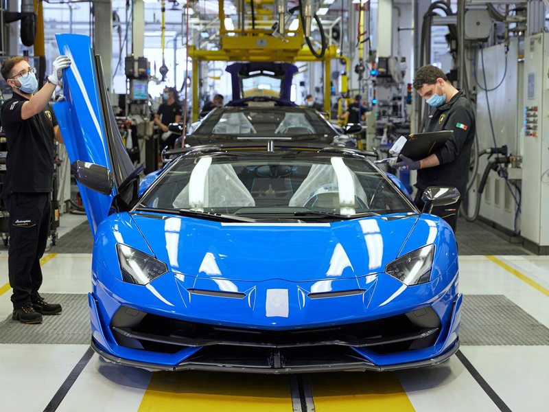 Masked Lamborghini factory workers assembling a blue Aventador SVJ Roadster with scissor door open on the production line
