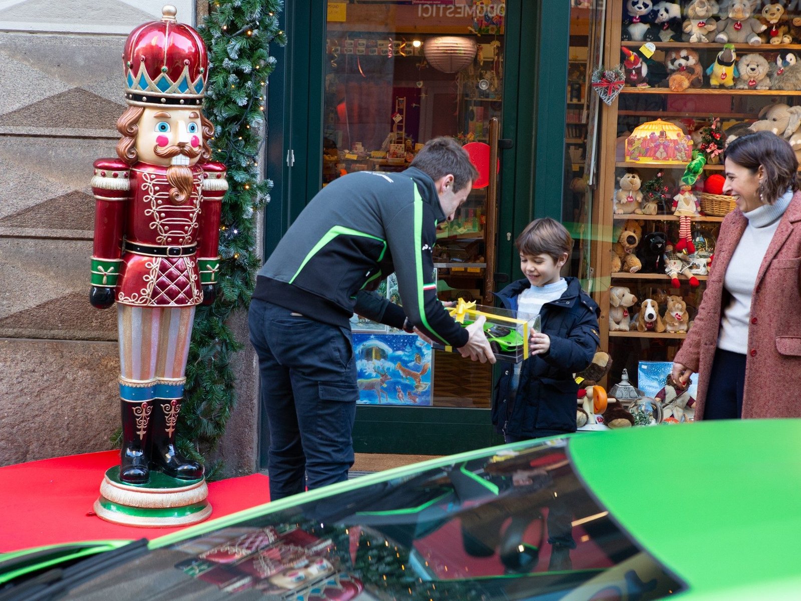 A man in a Lamborghini jacket hands a gift-wrapped toy car to a young boy outside a Christmas-decorated toy store, with a green Aventador SVJ visible in the foreground
