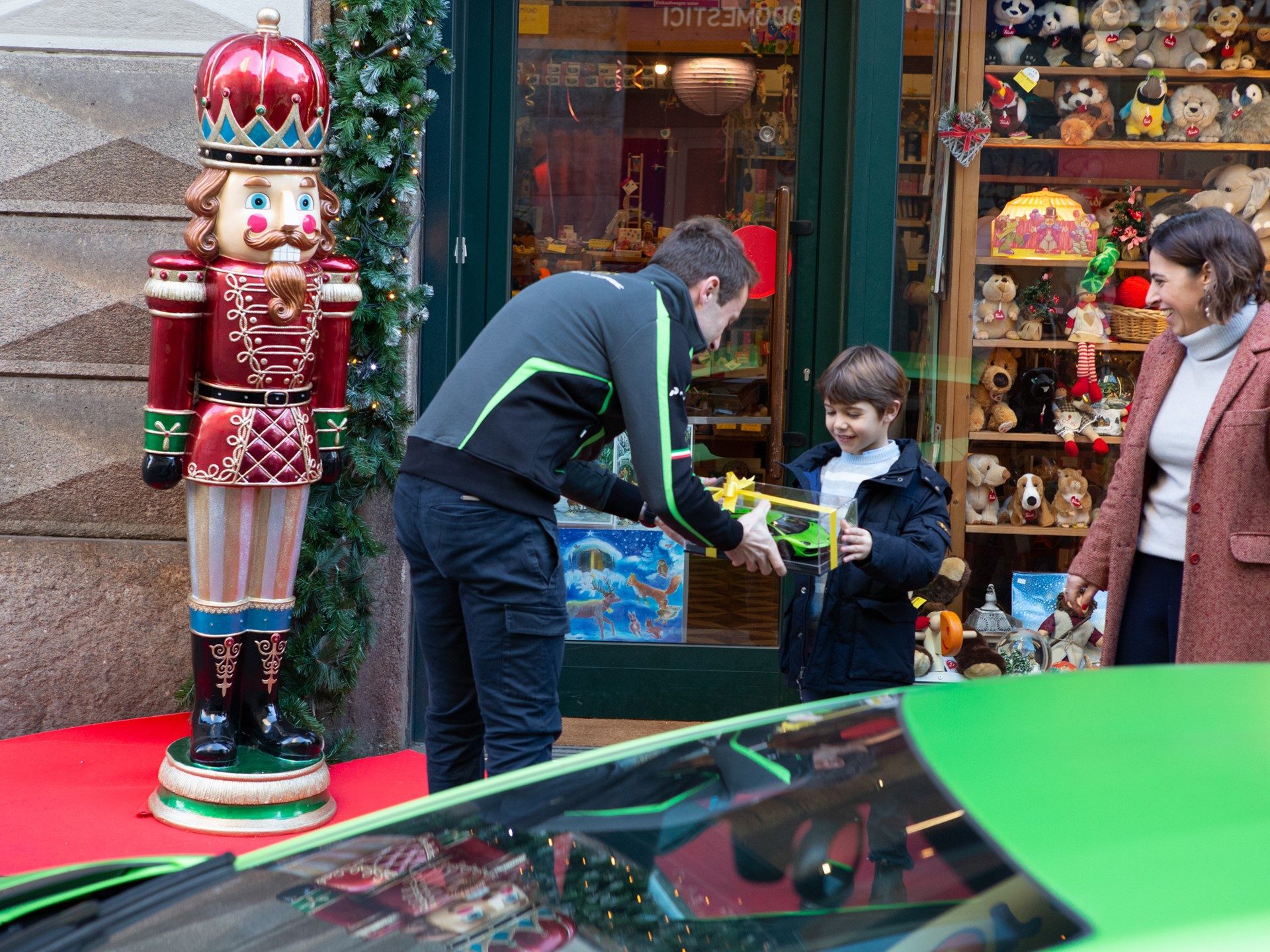 A man in a Lamborghini jacket hands a gift-wrapped toy car to a young boy outside a Christmas-decorated toy store, with a green Aventador SVJ visible in the foreground