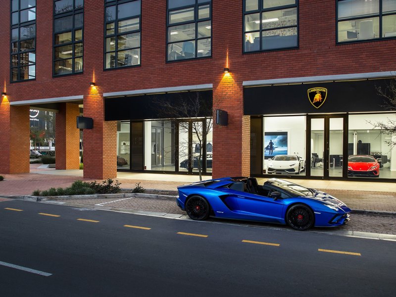 Blue Lamborghini Aventador S Roadster parked outside the new Lamborghini Cape Town showroom at night, with additional models visible through the brightly lit windows