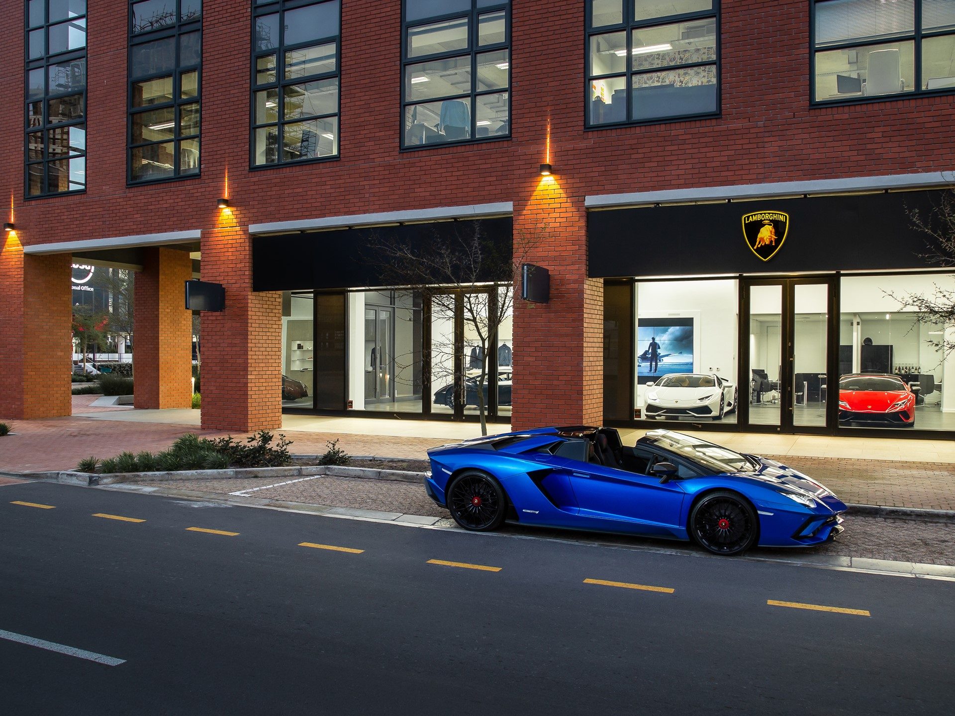 Blue Lamborghini Aventador S Roadster parked outside the new Lamborghini Cape Town showroom at night, with additional models visible through the brightly lit windows