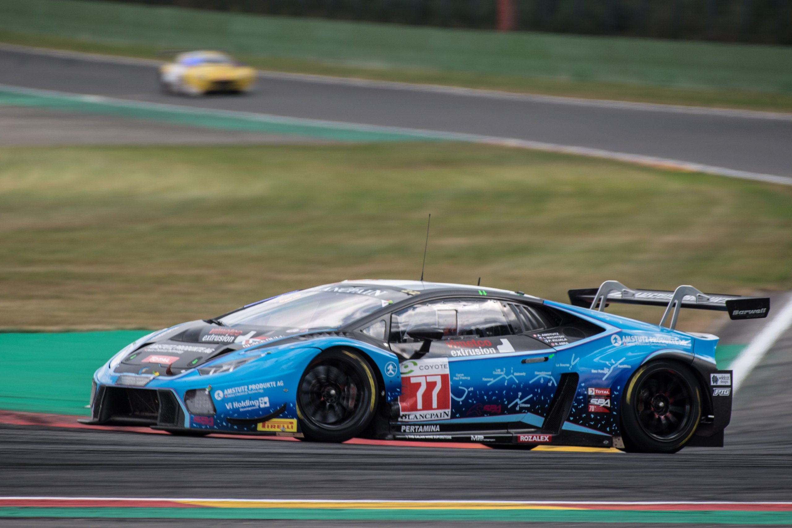 Blue Lamborghini Huracán GT3 with sponsor livery racing at Spa-Francorchamps with a blurred competitor visible behind