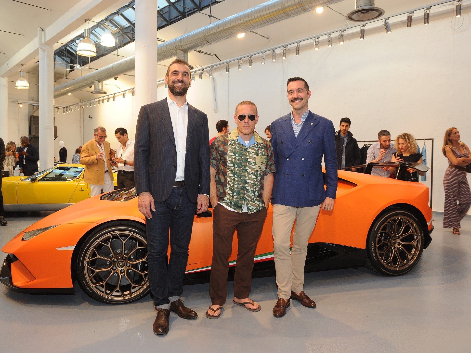 Guests gathered around the orange Lamborghini Huracán Performante at the Milan fashion collection event, with a yellow Lamborghini visible in the background