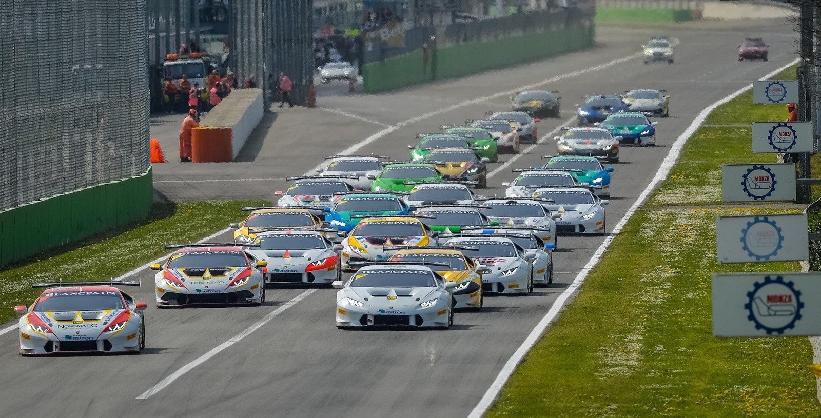 A packed grid of Lamborghini Huracán LP 620-2 Super Trofeo race cars in multi-colored liveries lined up on a circuit, viewed from an elevated front angle with spectators and officials in the background.