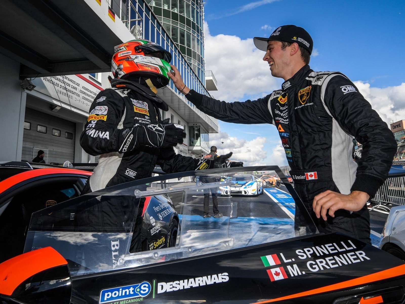 Two Lamborghini Super Trofeo drivers interact beside a black and orange Huracán Super Trofeo Evo on the Nürburgring grid