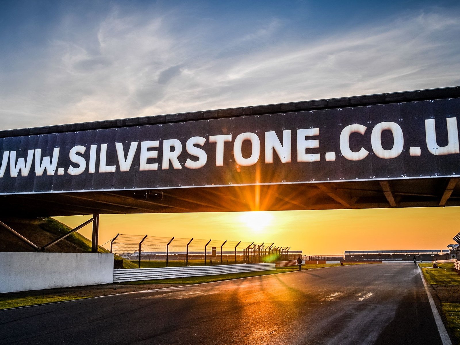Silverstone circuit at sunset with the track stretching toward the horizon under an orange sky, a bridge overhead displaying the Silverstone URL