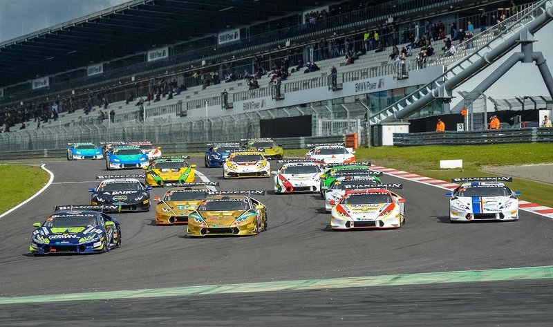 Multiple Lamborghini Huracán Super Trofeo race cars in colorful liveries battling for position on a racetrack with spectators in the grandstands