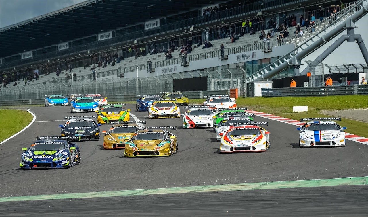 Multiple Lamborghini Huracán Super Trofeo race cars in colorful liveries battling for position on a racetrack with spectators in the grandstands