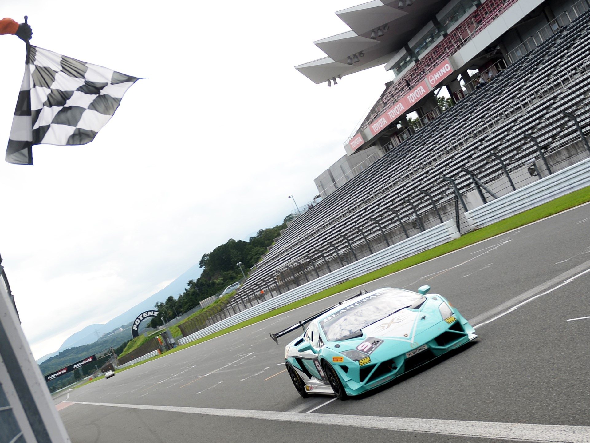 A light blue Lamborghini Gallardo Super Trofeo race car crosses the finish line under a waving checkered flag at a racetrack