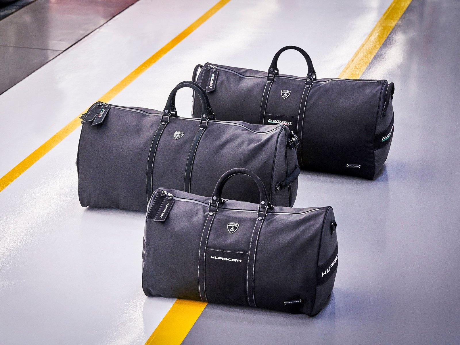 Three grey Lamborghini Tecknomonster soft bags with Huracán, Aventador, and Urus model names and Lamborghini shield logos visible