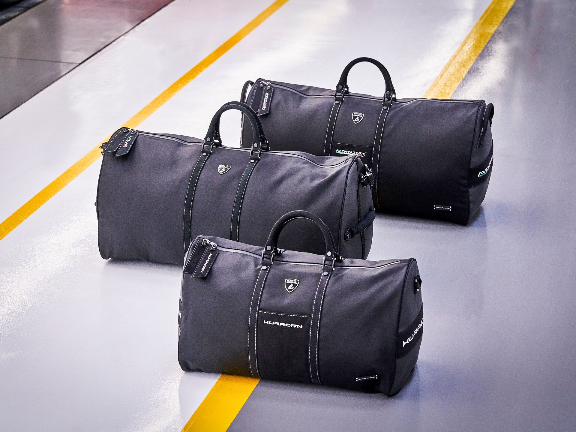 Three grey Lamborghini Tecknomonster soft bags with Huracán, Aventador, and Urus model names and Lamborghini shield logos visible