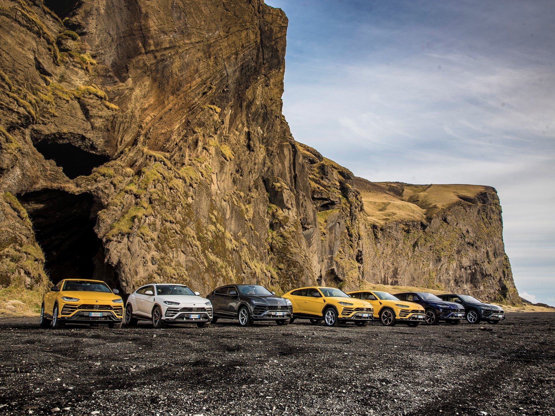 Multiple Lamborghini Urus SUVs in yellow, white, grey, and blue lined up on Iceland's black sand beach with rugged cliffs behind them